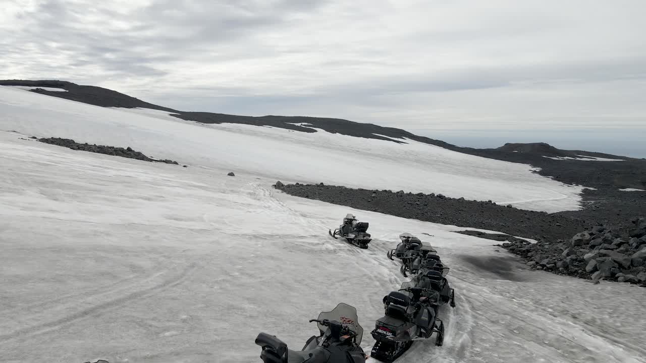 A drone sweeps across Mount Katla's snowy expanse in South Iceland, tracking a convoy of snowmobiles carving through the pristine white, embodying the thrill of wilderness exploration