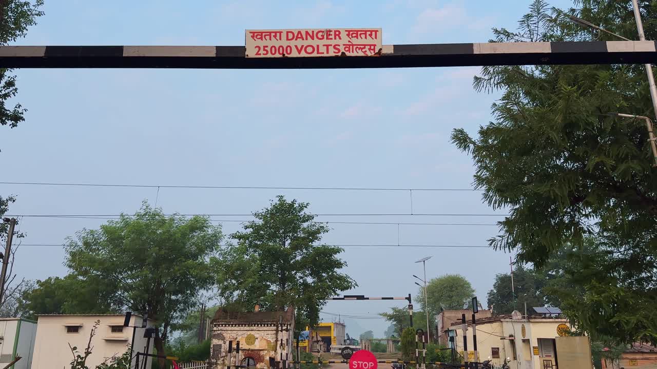 Tilt-up shot starting from the dusty road leading toward a closed railway crossing, rising to reveal the overhead danger sign,