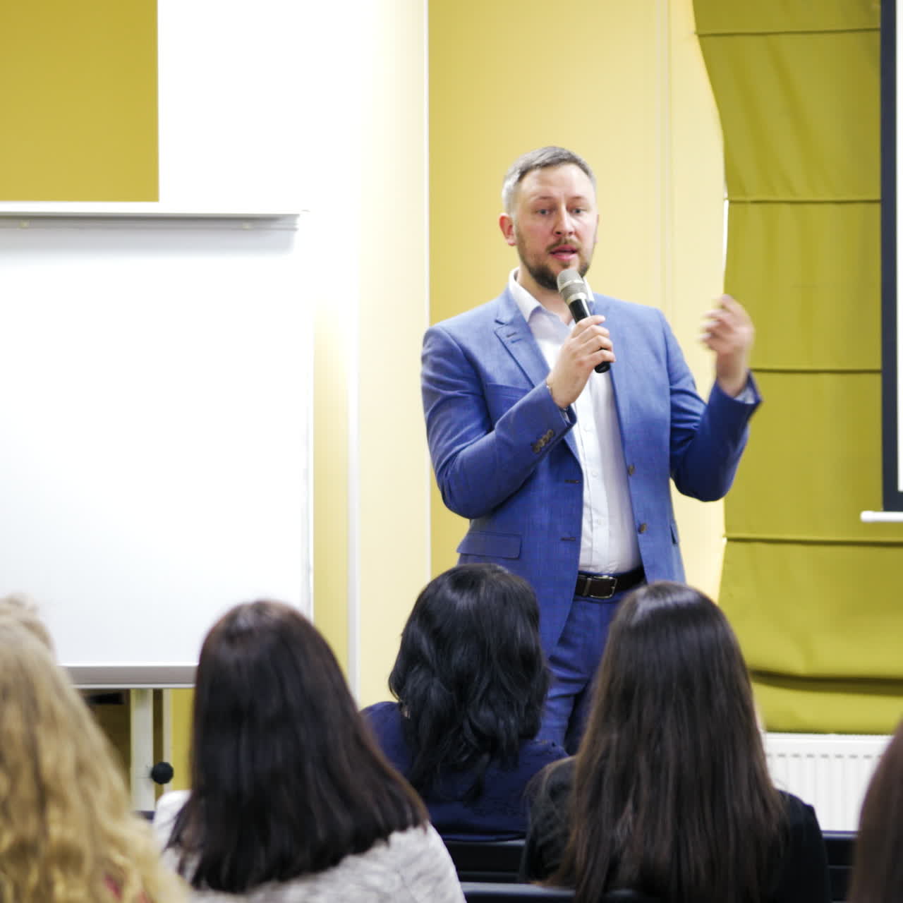Businessman explains new program to the audience. Handsome man speaker conducts a lecture while standing near the white board in a conference hall.