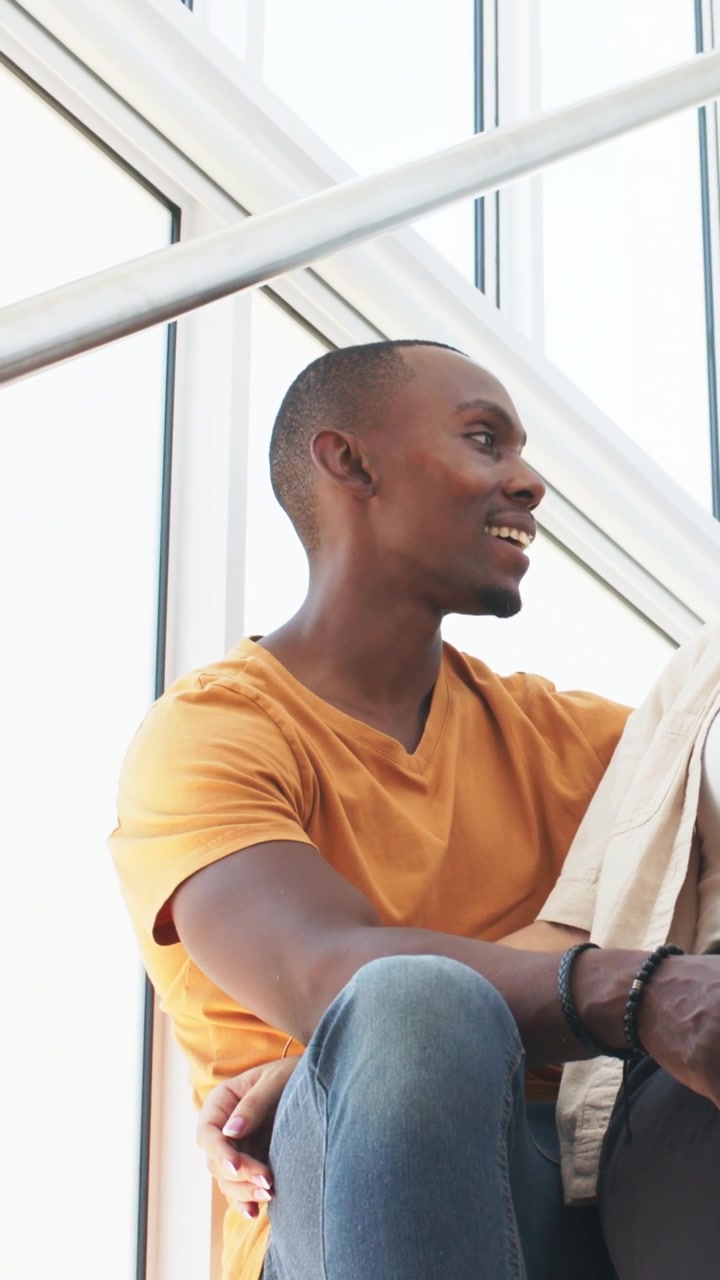 Vertical video: Smiling man in mustard tee leaning toward her on stair by window, replying to her