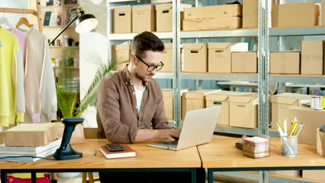 caucasian designer man in eyeglasses working on laptop while sitting at desk in clothing shop warehouse