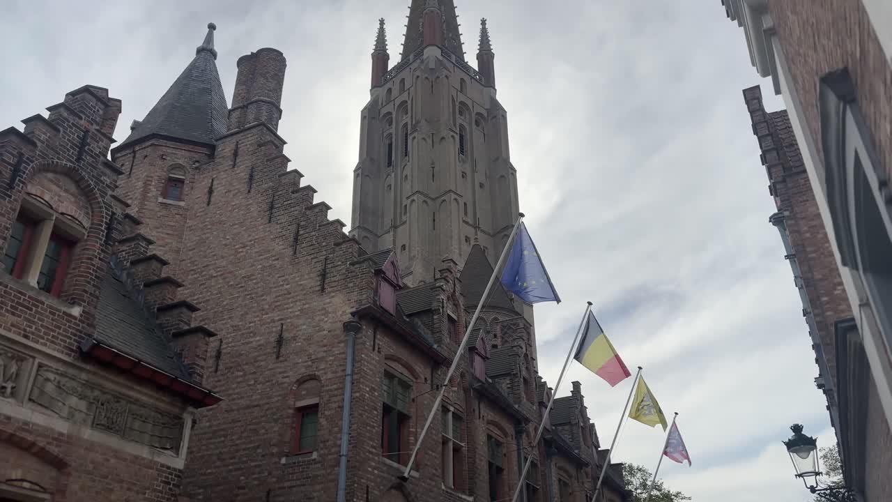 Low angle shot of a medieval church in Bruges with Belgian and Flemish flags waving under cloudy sky