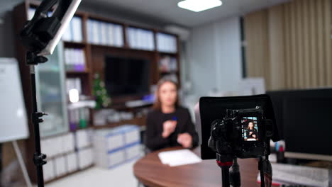 camera on tripod records a female caucasian woman. female blogger talks and gestures with her hands. blurred backdrop.