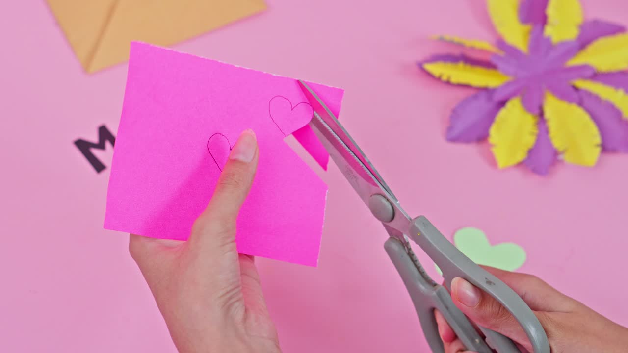 Hands using scissors to cut out heart shapes from pink paper for Mother's Day craft project