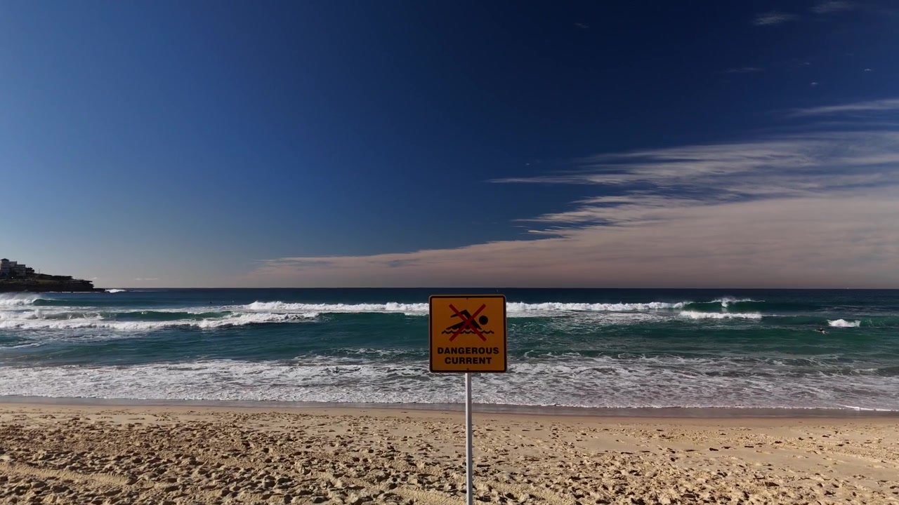 Placa de "Proibido nadar, corrente perigosa" na praia em um dia ensolarado perfeito com ondas e céu azul e prancha de resgate. A câmera se move de uma tomada ampla para um close da placa.