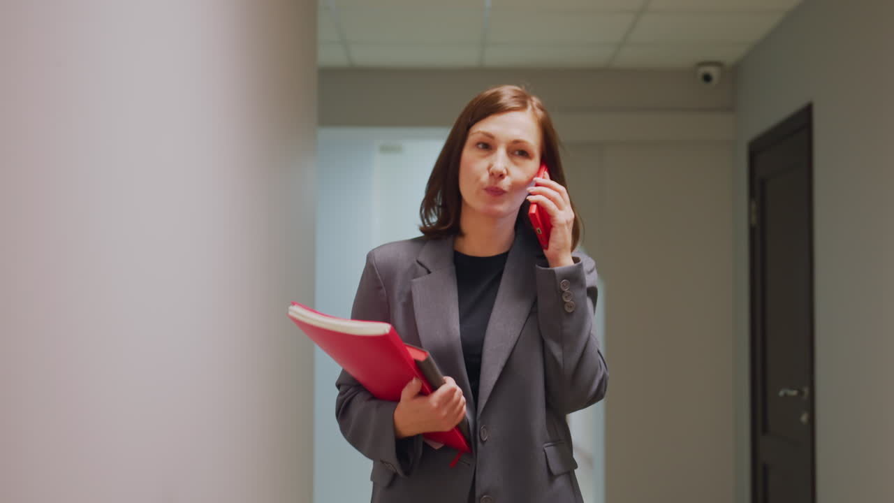 Professional businesswoman walking in office corridor while talking on phone and holding red folder. Confident and engaged communication in modern, minimalist office environment