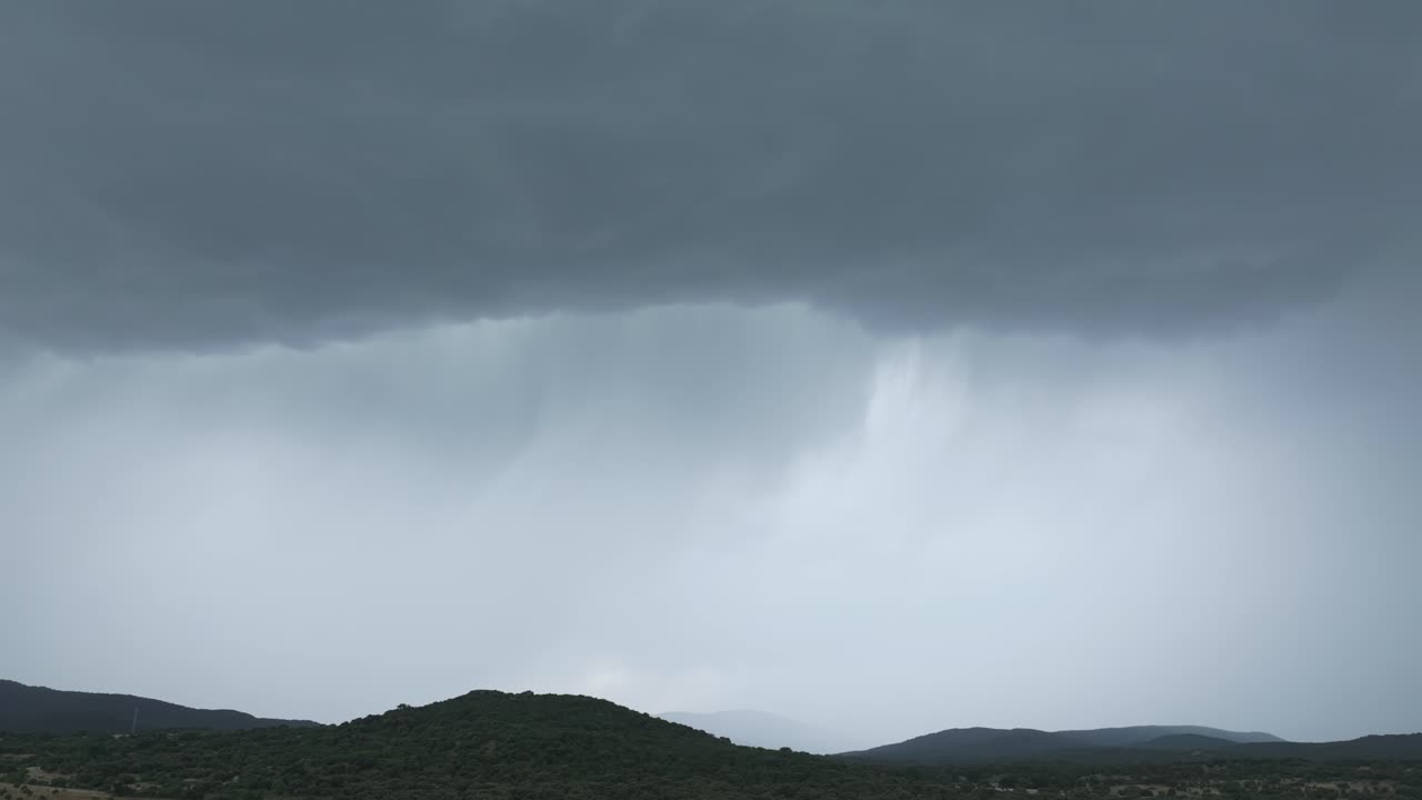 Drone shot panning right across a sky filled with dark storm clouds over shaded hills. Rain descends in distant streaks, bringing life-giving water to the land beneath a dramatic, powerful sky