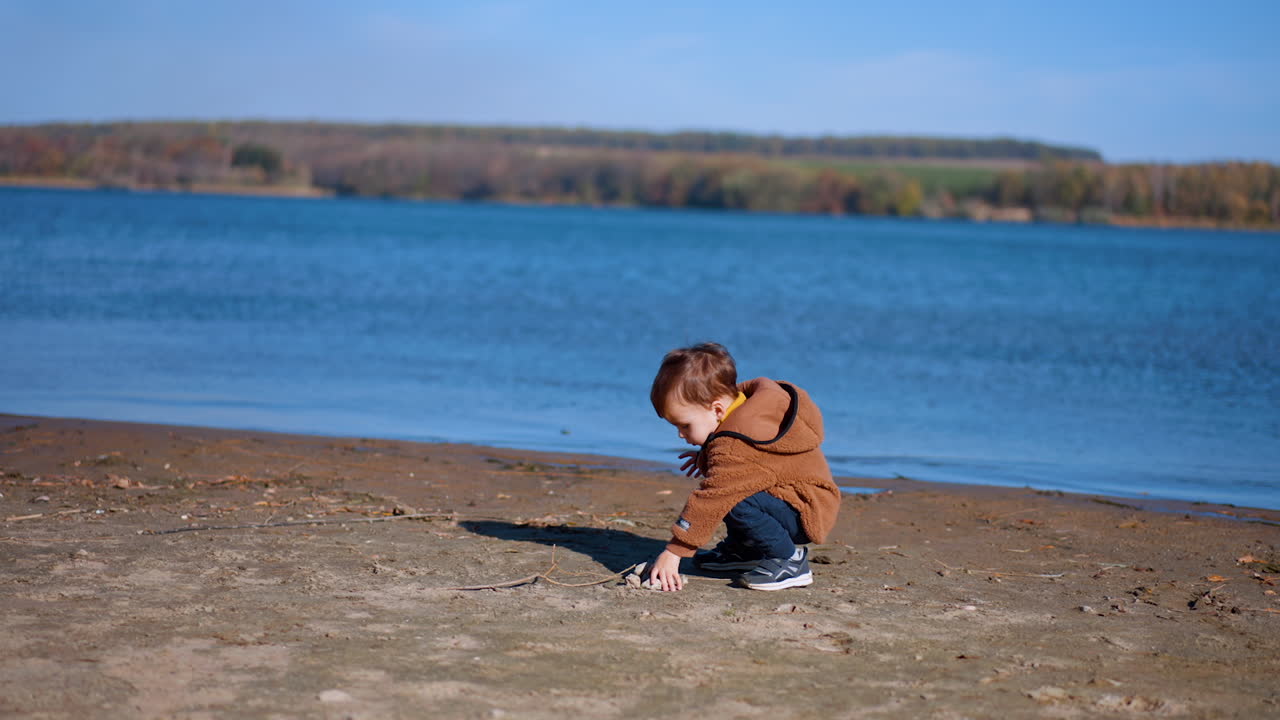 Eighteen months old toddler walking by the river bank. Baby boy picks up the stones and throws them into water.