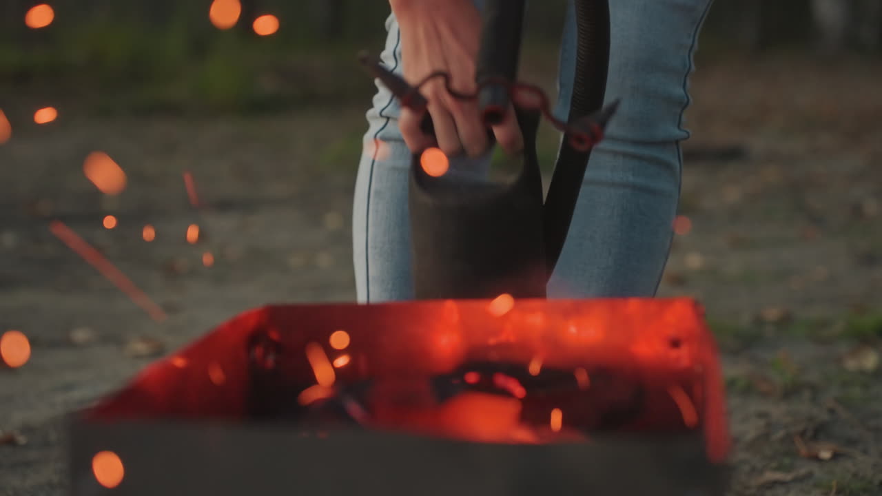 Campeggiatore che versa liquido su braci ardenti, gambe in jeans e mani che tengono un bollitore di metallo con cinghia, scintille che si alzano da un braciere rettangolare, manutenzione serale del campeggio e azione di sicurezza catturate.