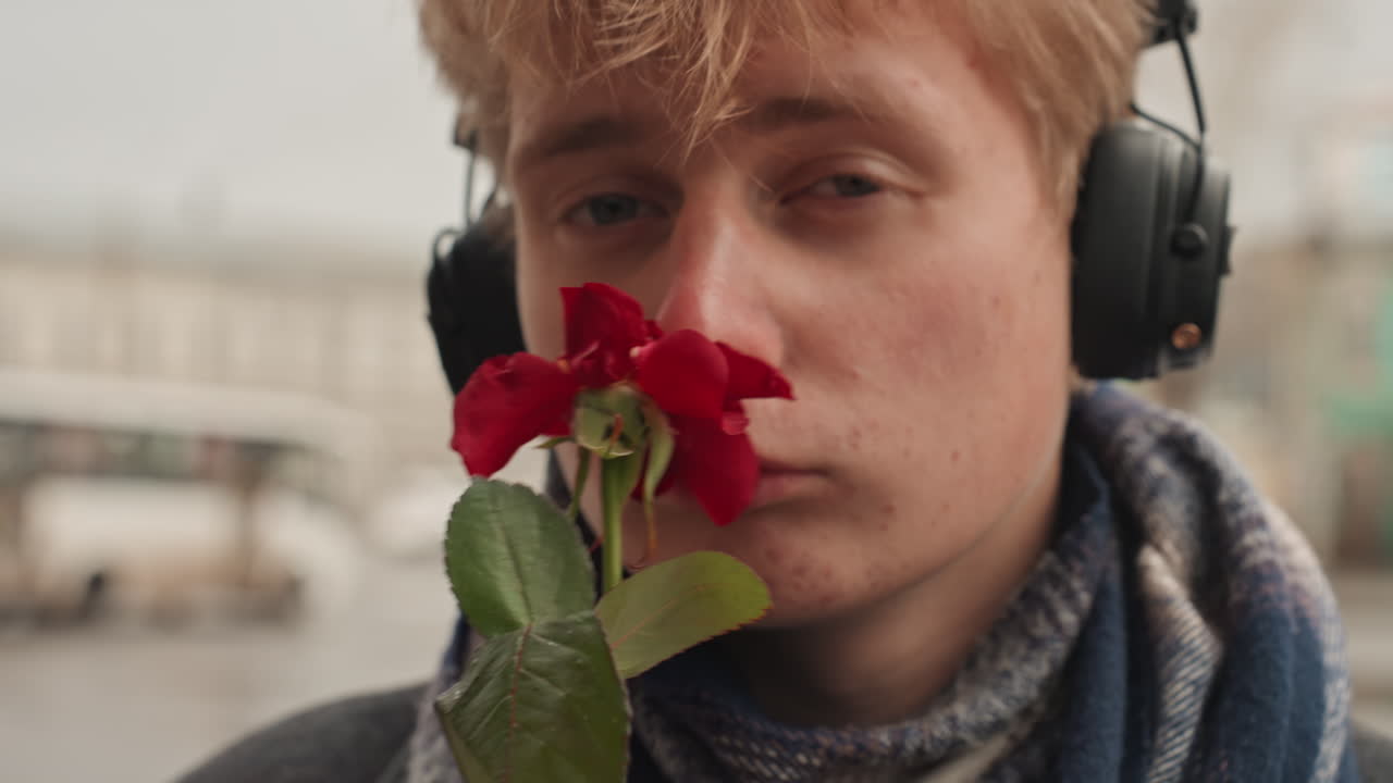Nahaufnahme eines jungen weißen Kreativen mit roter Rose, Kopfhörer umrahmen das Gesicht und nachdenklicher Blick, verletzlicher Ausdruck mit strukturierter Haut und Sommersprossen, geringe Tiefenschärfe auf Blütenblättern und verschwommener urbaner Brückenhintergrund