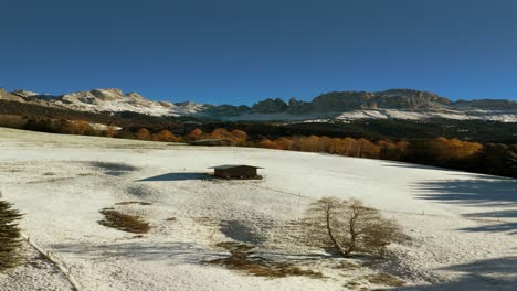 Vertical drone footage of a snowy alpine meadow with a wooden hut and vibrant autumn trees below the Rosengarten mountains in the Dolomites of South Tyrol, northern Italy