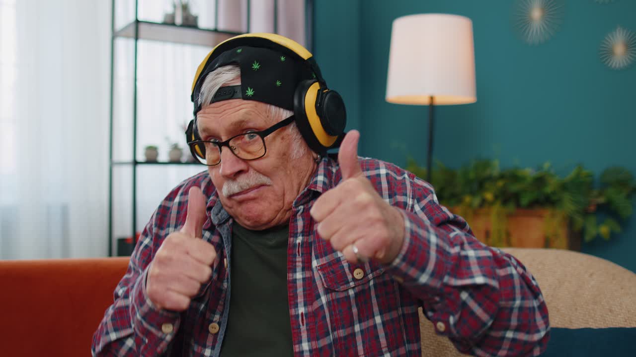 Un anziano felice con le cuffie wireless che balla e canta su un comodo divano nel soggiorno di casa