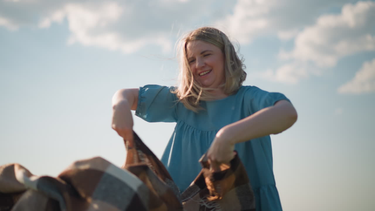 A woman smiles slightly as she holds a plaid scarf, with the wind gently blowing it in a serene outdoor setting. The scene captures a joyful and carefree moment under a bright sky