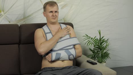 painful, bored man with a broken arm wearing arm brace sitting on a sofa
