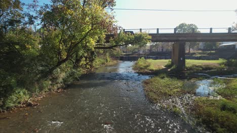 Aerial-pass-under-bridge-following-creek-approaching-waterfall-in-Old-Town-Helena,-Alabama