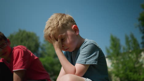 a tired-looking little boy wearing a blue shirt rubs his face with his left hand, with a blurred view of his elder brother looking at him