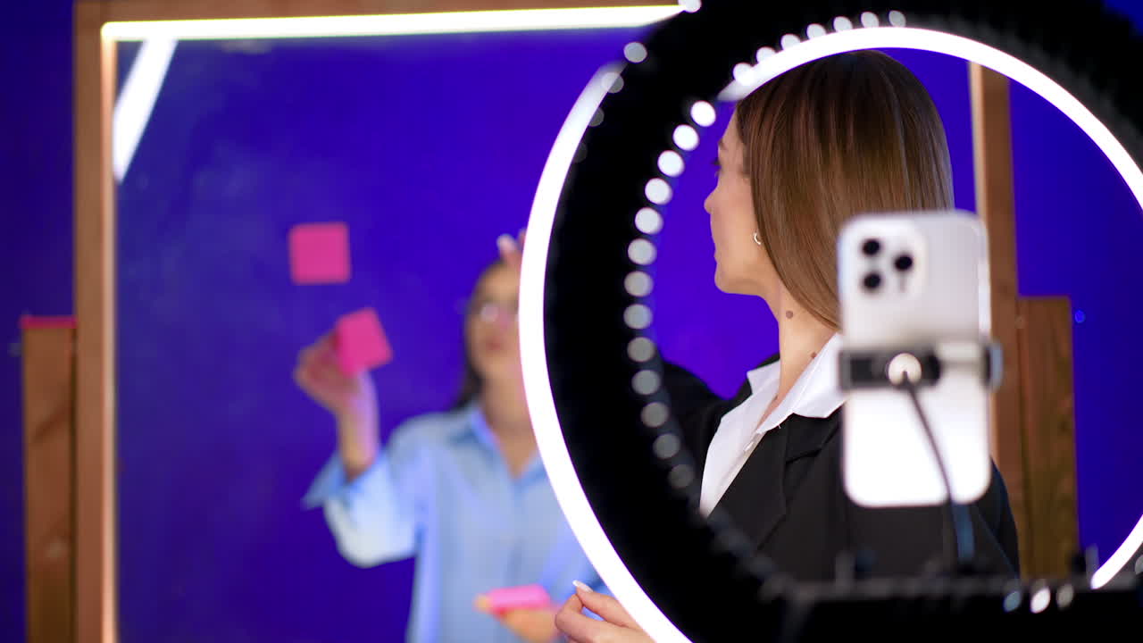 Beautiful Caucasian woman with long hair stands looking at camera with light ring. Blogger talks passionately gesturing with her hands. Woman turns around to point at glass wall behind which a girl stands.
