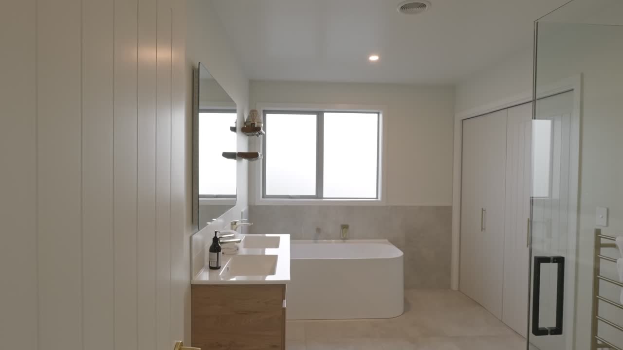 Acercamiento en cámara lenta a un baño blanco luminoso con bañera exenta, mueble de baño de madera, espejo redondo y mampara de ducha de cristal con luz natural de ventana