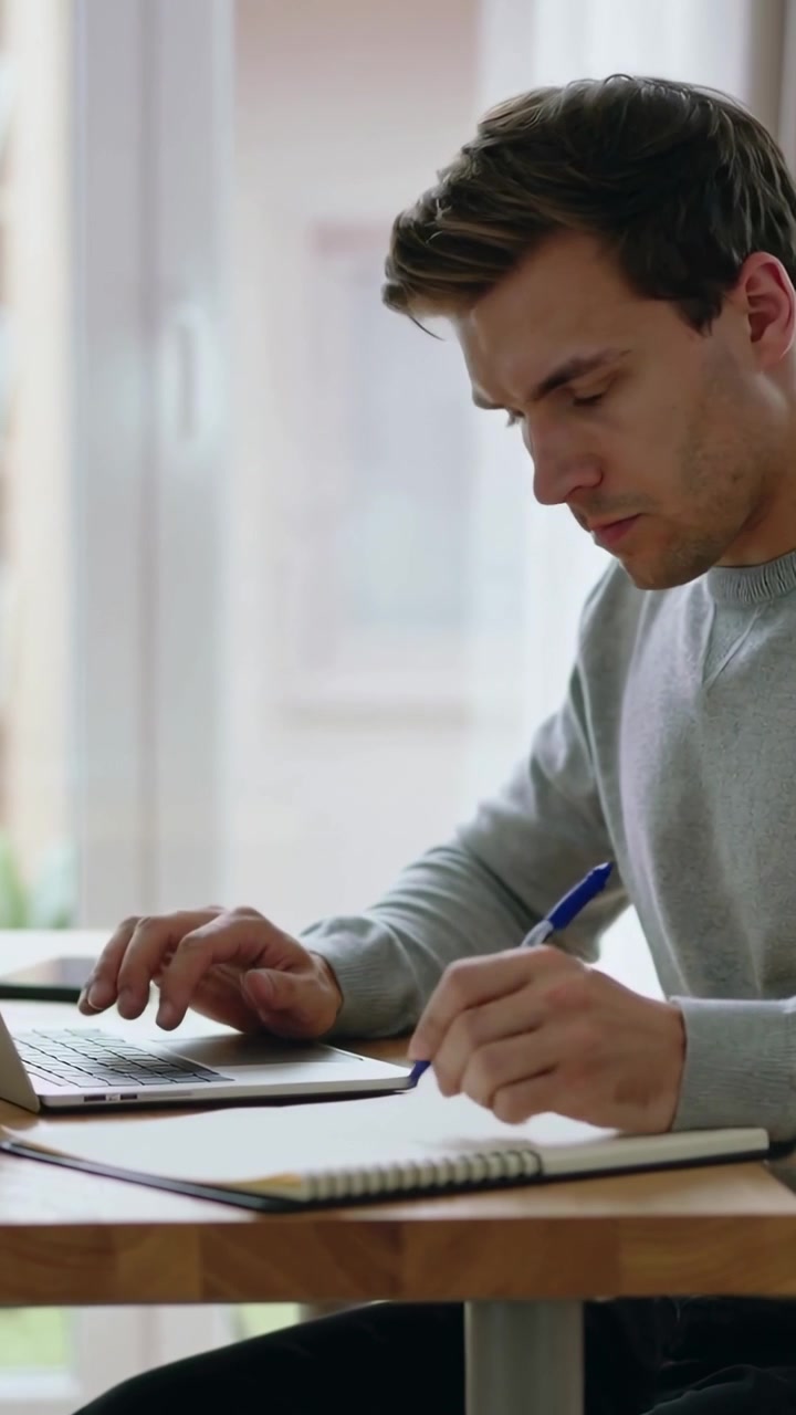 Vertikal video: Leser på laptop, mann i genser skifter mellom å skrive på tastatur og skrive for hånd hjemme med notater og penn