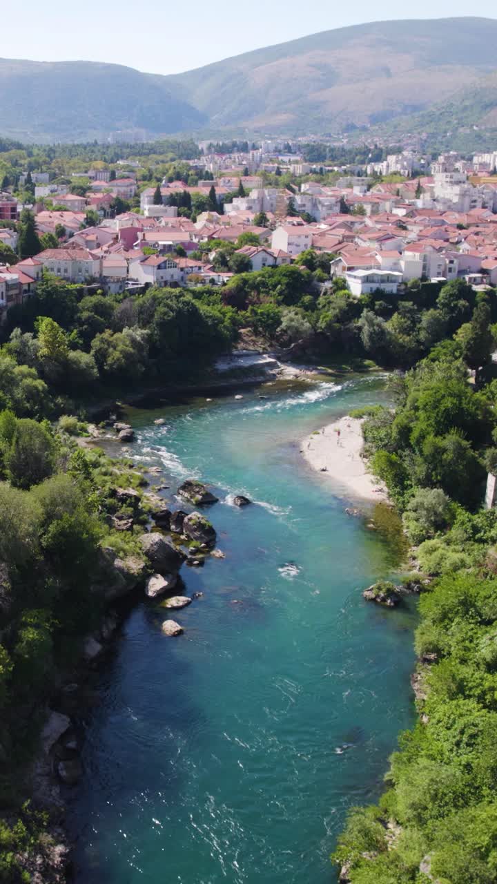 Aerial view of Neretva river flowing through Mostar city in Bosnia and Herzegovina, surrounded by houses and mountains. Vertical Video