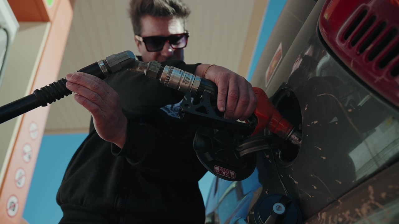 Man in glasses inserting red fuel nozzle into muddy off road vehicle at petrol station