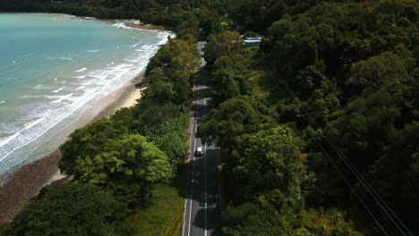 modern silver car driving on a road along the seaside coast with sandy beach and lush green tree forest and blue water sea