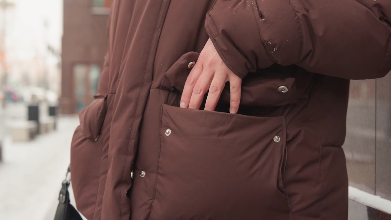 Waist-level view of person wearing brown quilted puffer coat with hands near front pockets, visible hand movement suggesting reaching into or adjusting pocket; snowflakes and water droplets cling to