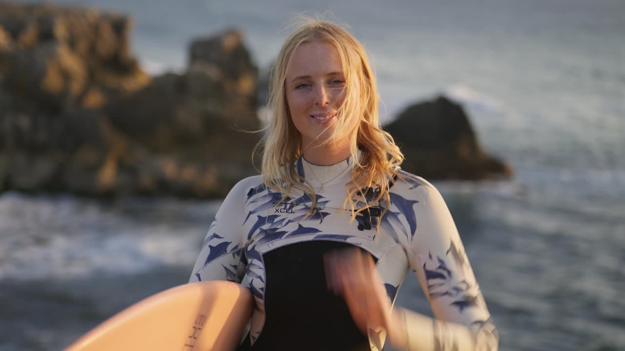4k cámara lenta retrato de cerca de una joven surfista sonriente con el pelo rubio soplando en el viento mirando la puesta de sol