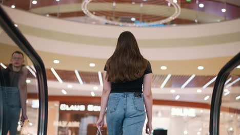 back view of young lady with shopping bags stepping off escalator in mall, boy passing by in background, vibrant lights and reflections from decorative ceiling overhead