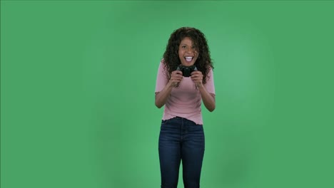 retrato de una hermosa joven afroamericana está mirando a la cámara y jugando a un videojuego usando un controlador inalámbrico con alegría y emociones de wow. bruneta ardiente con cabello ondulado en vaqueros y una blusa beige en una pantalla verde en el estudio