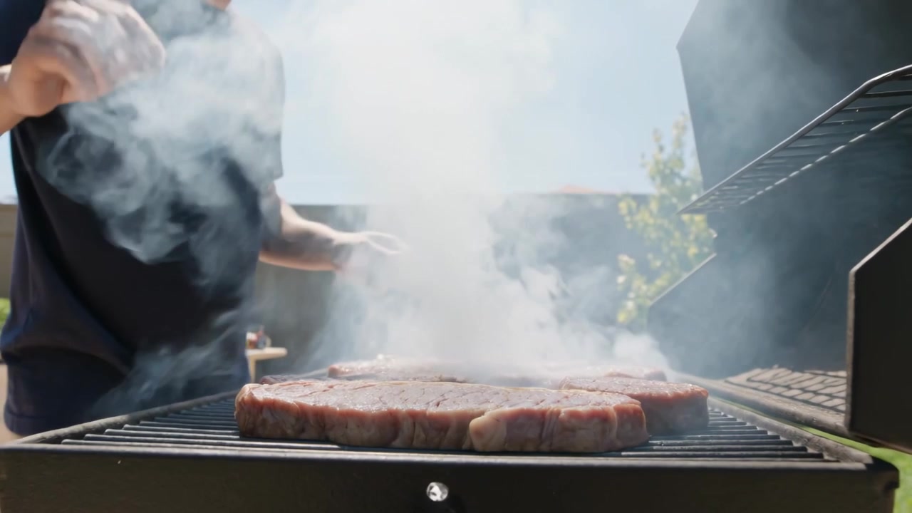 Kok i marineblå T-shirt nærmer sig og lægger bøffer på havegrillen med en tang for at svitse dem midt i røgen.
