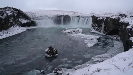 Vista-Aérea-De-La-Cascada-Godafoss-Que-Desemboca-En-El-Río-Skjalfandafljot-En-La-Isla-Norte-Durante-El-Invierno---Flujo-De-Hielo-Y-Iceberg-Flotando-En-La-Superficie-Del-Agua