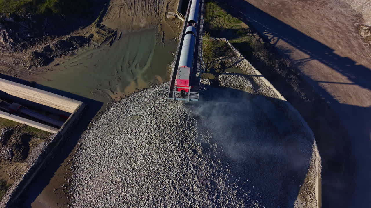 Heavy machinery transfers gravel to a large mound at a construction site during bright daylight, showcasing industrial activity and earth movement
