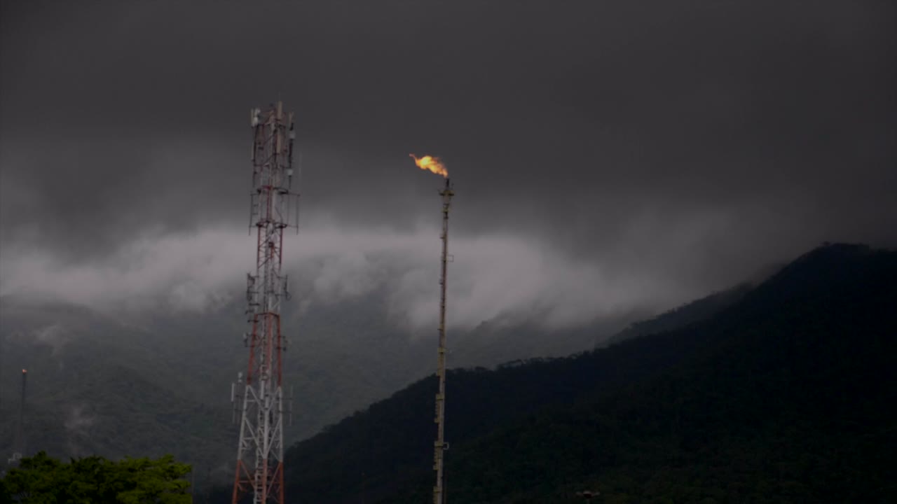 toma panorámica a través de la torre de combustión de gas mientras el mástil pasa por el cielo oscuro - ladera de la montaña en el fondo
