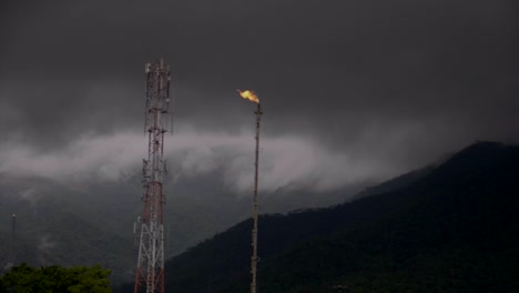 Panning-shot-across-gas-burn-off-tower-as-mast-passes-across-dark-sky---mountain-slope-in-background