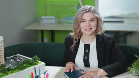 professional woman closes book with gentle smile while seated in modern office with green decor, desk accessories, architectural model, and laptop, radiating confidence, calmness, and productivity