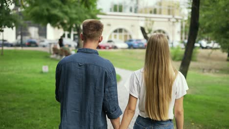 una joven pareja positiva camina tomados de la mano, se reúnen en un parque de la ciudad de verano. la niña y el niño son rubios. caminata feliz en el parque. vista posterior