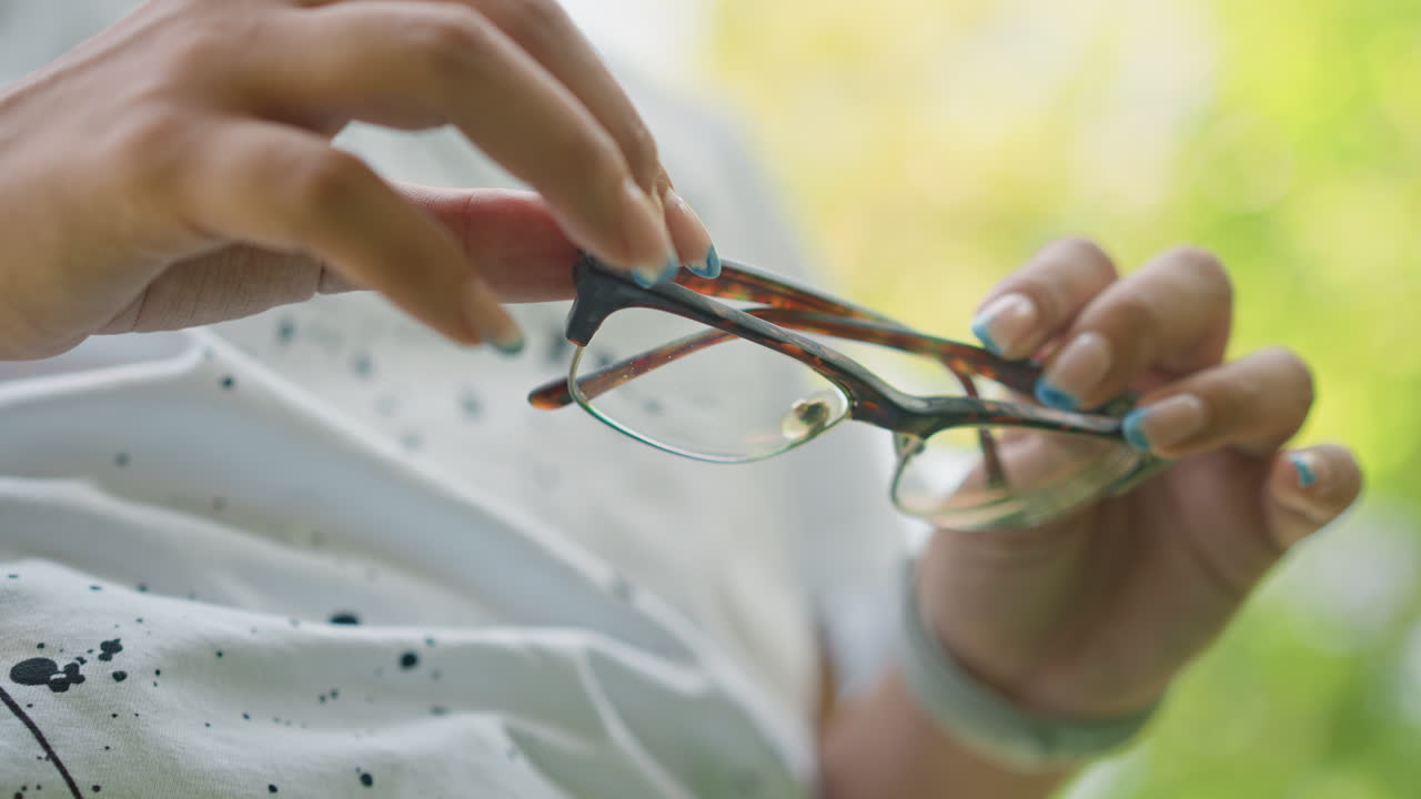 Primer plano de unas manos sujetando unas gafas al aire libre a la luz del sol. Escena tranquila y delicada de una persona ajustando una montura de carey con manicura turquesa, camisa blanca moteada, fondo suave con efecto bokeh y enfoque táctil.