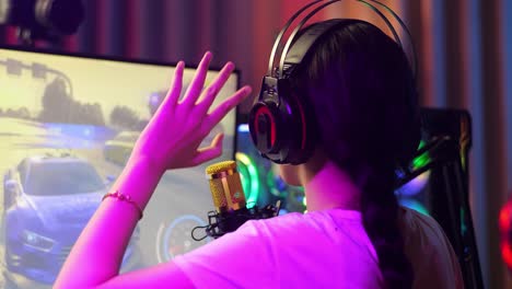 close up back view of asian girl streamer playing car racing game over network on computer, talking on the microphone.  live stream video game, desk illuminated by rgb led strip light