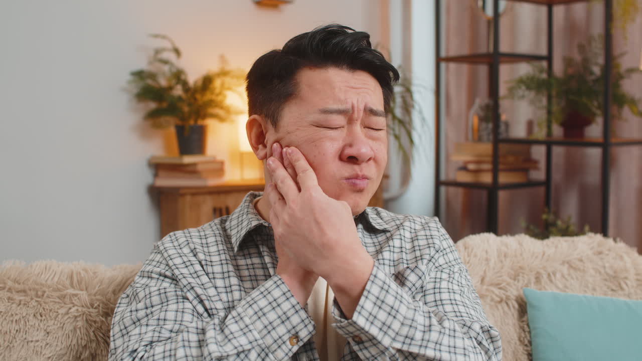 Young man touching cheek with expression of terrible suffer from painful toothache on sofa at home