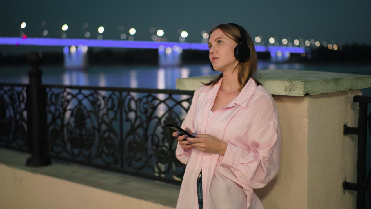 chic blanco en top rosa y auriculares descansando en la valla, perdido en sus pensamientos con las luces del puente iluminadas reflejadas en el río en la distancia, y la vista borrosa de los coches que se mueven en el puente