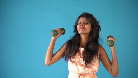A-young-Indian-girl-in-orange-frock-standing-with-dumbbells-and-dancing-in-an-isolated-blue-background