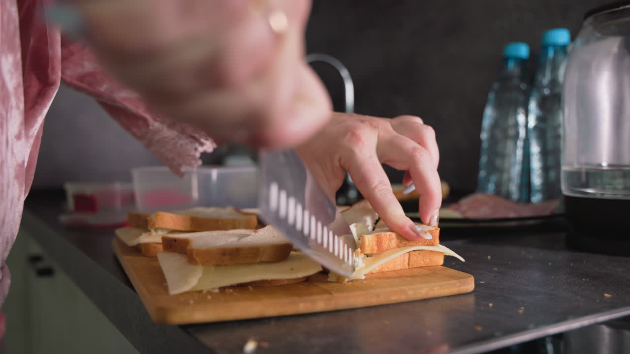 Caregiver slices cheese sandwich on wooden board with sharp knife while preparing school lunch in modern kitchen, showcasing close-up hands, layered ingredients, and focused food preparation process