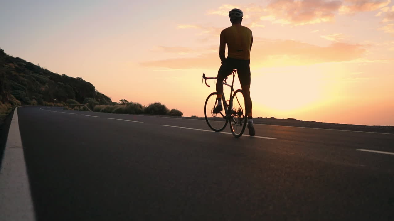 A youthful athlete, dressed in a yellow t-shirt, and sports gear, sits atop a bike on a mountain peak, gazing at the exquisite mountains and the descending sun. Moments of repose follow training