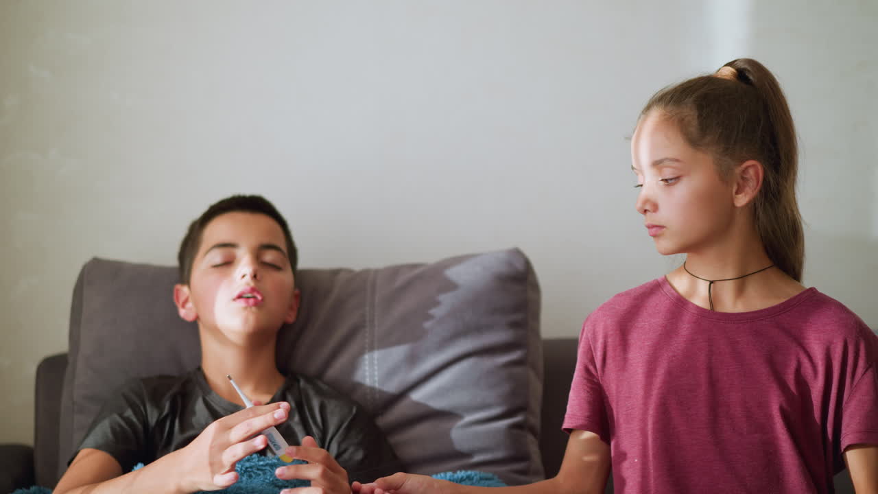 Sick boy sitting on couch collects thermometer from sister to check his temperature, placing it under his armpit while covered with a cozy blanket, soft natural light filters through the window