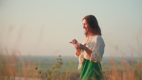 fair skinned woman in flowing green dress standing cliff edge overlooking river valley, blowing iridescent soap bubble into warm sunset sky, tall grasses swaying capturing peaceful moment in nature