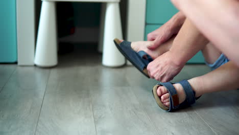 un niño pequeño viste zapatos solo, el niño se sienta en el suelo y se pone sandalias azules, la madre ayuda a vestirse