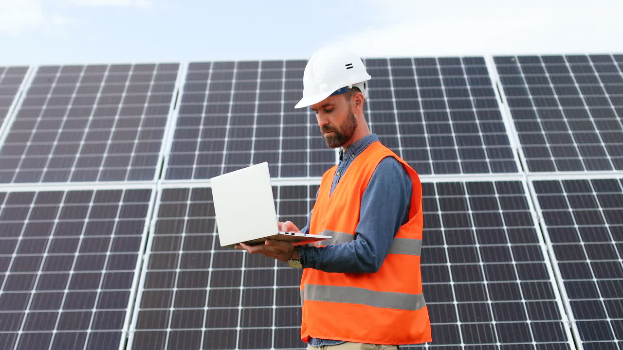 Young male engineer with helmet and vest near solar panels using laptop