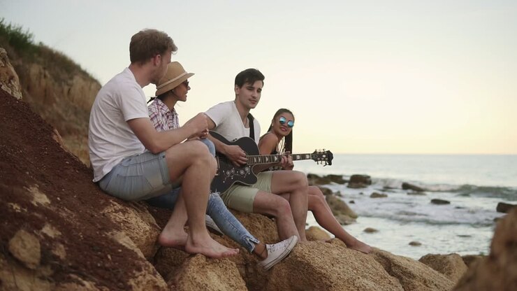gruppo di giovani amici hipster seduti sulle rocce in riva al mare e suonando la chitarra e cantando canzoni