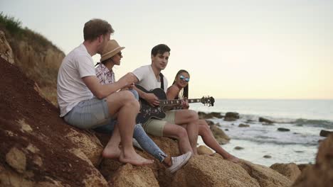Gruppe-Junger-Hipster-Freunde,-Die-Auf-Den-Felsen-An-Der-Küste-Sitzen-Und-Gitarre-Spielen-Und-Lieder-Singen